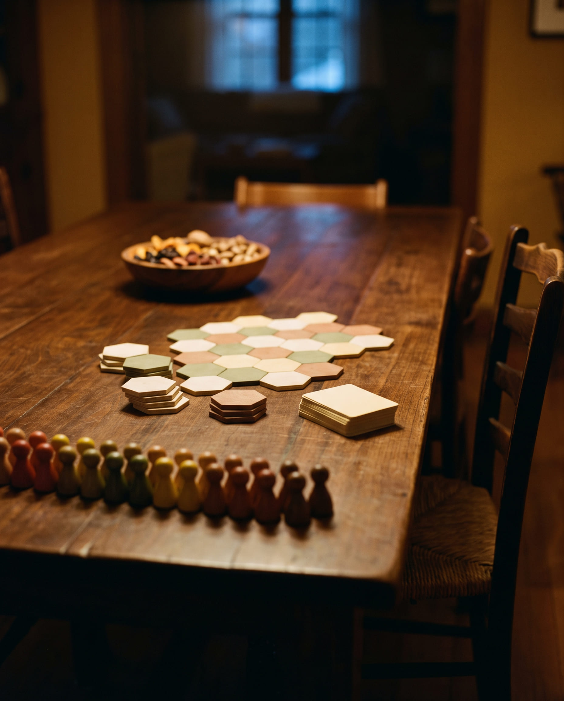 A dark walnut table before anybody sits down: a row of turned wooden player pieces laid out in colour order along the near edge, a part-built hex board in the middle with the rest of the tiles still stacked beside it, a bowl of snacks pushed to the far end and a rush-seat chair drawn in at the right.