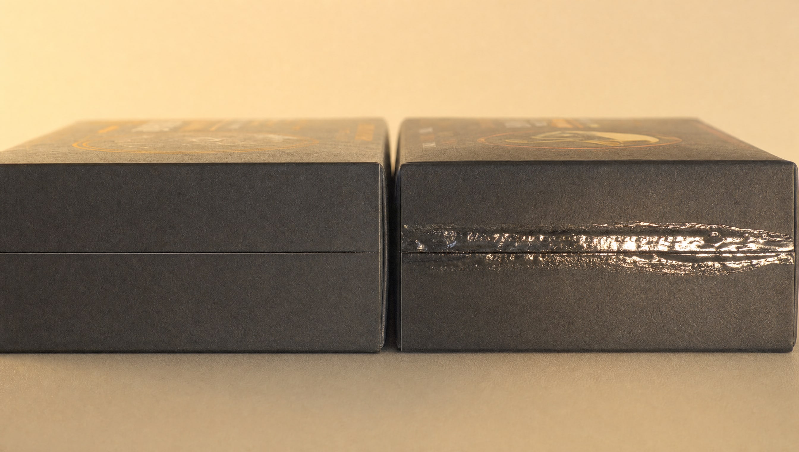 Two identical sealed blind boxes side by side under hard raking light, the right-hand flap showing a glossy band of dried glue where it was opened and stuck back down.