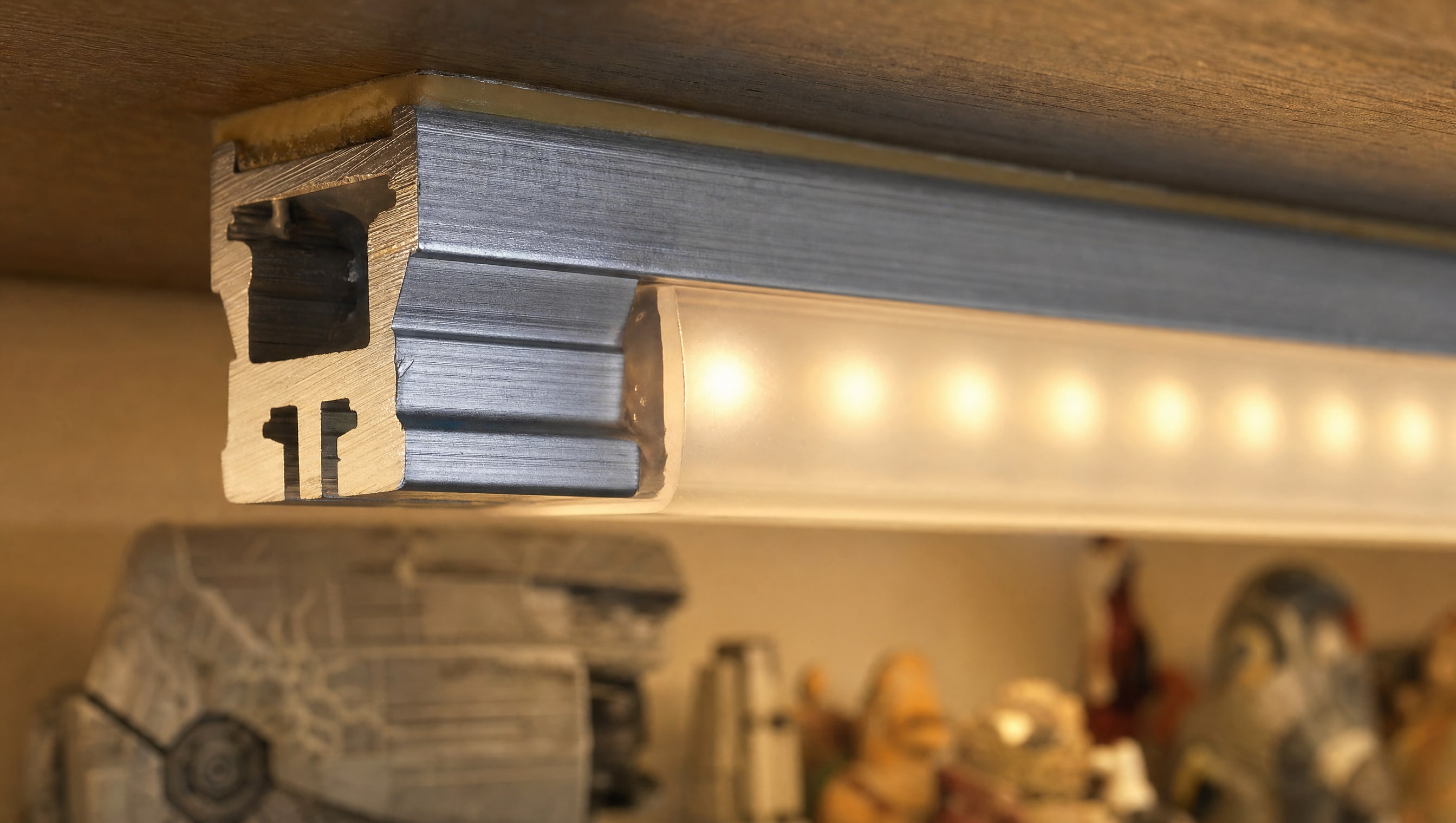 Macro on the end of an anodised aluminium LED channel under a shelf, the frosted diffuser seated in its groove and warm light spilling onto the deck.