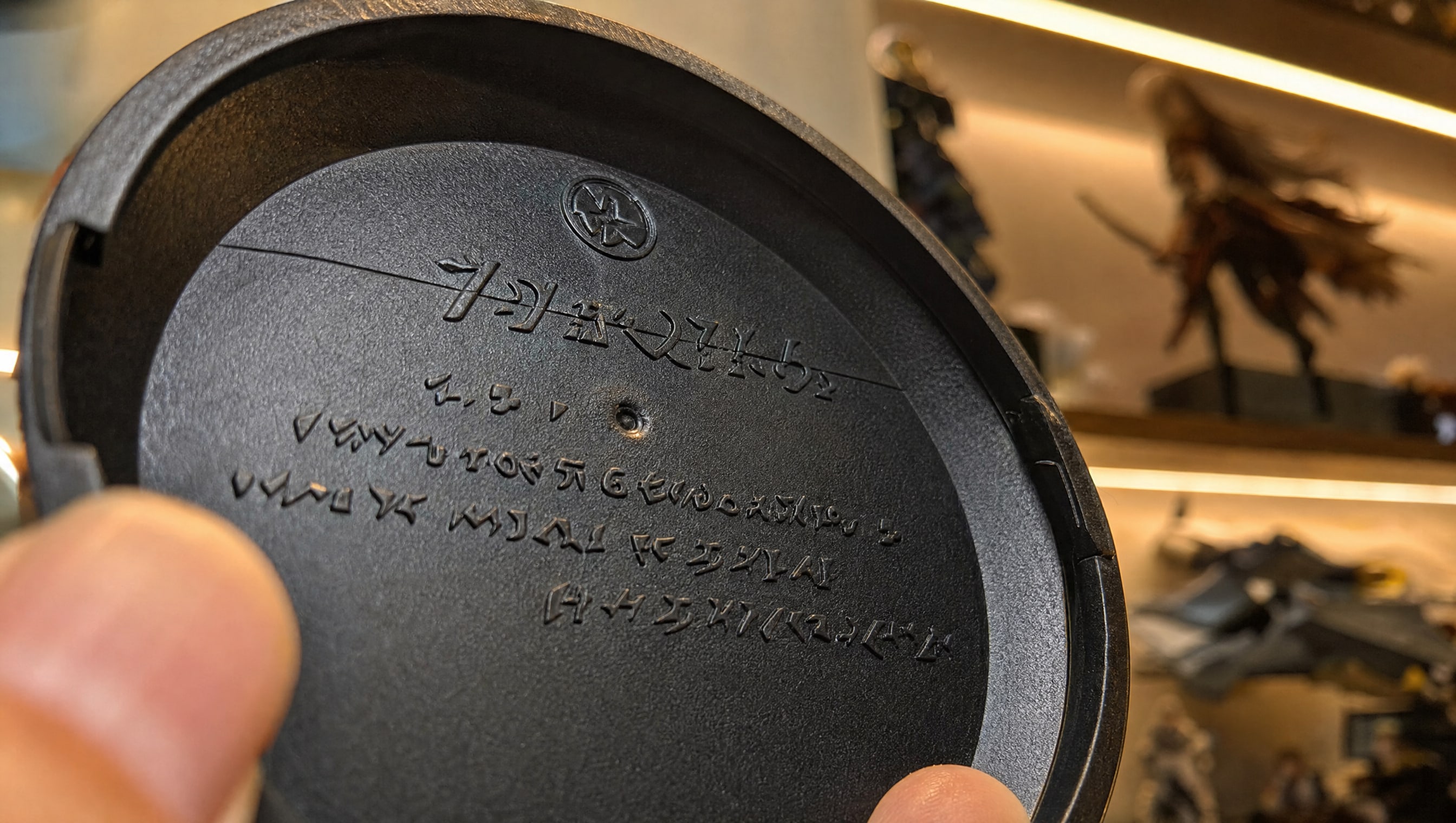 Macro on the underside rim of an invented anime figure’s display base, moulded maker and licence lettering catching warm shelf light.