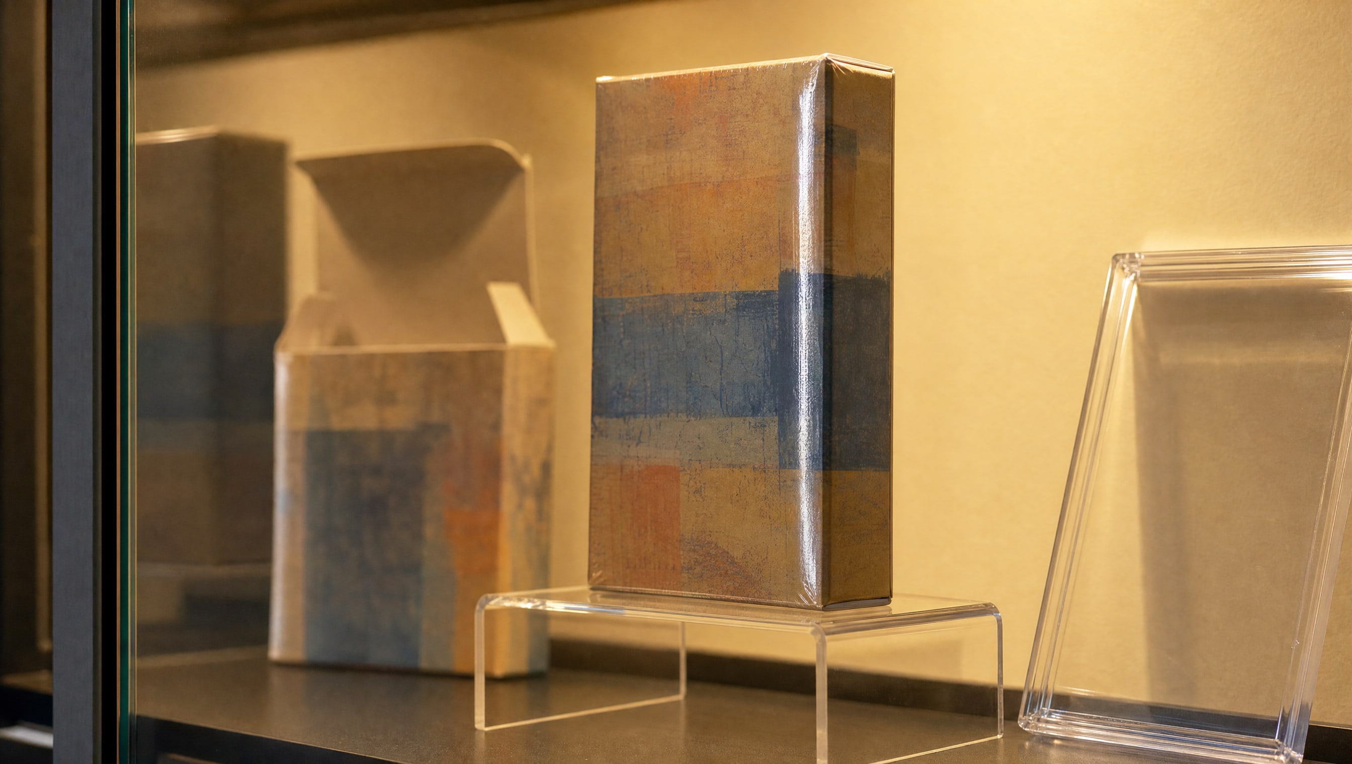 A sealed invented boxed game standing on a riser inside a case, warm LED light across the film, a loose copy of the same box lying open behind it.