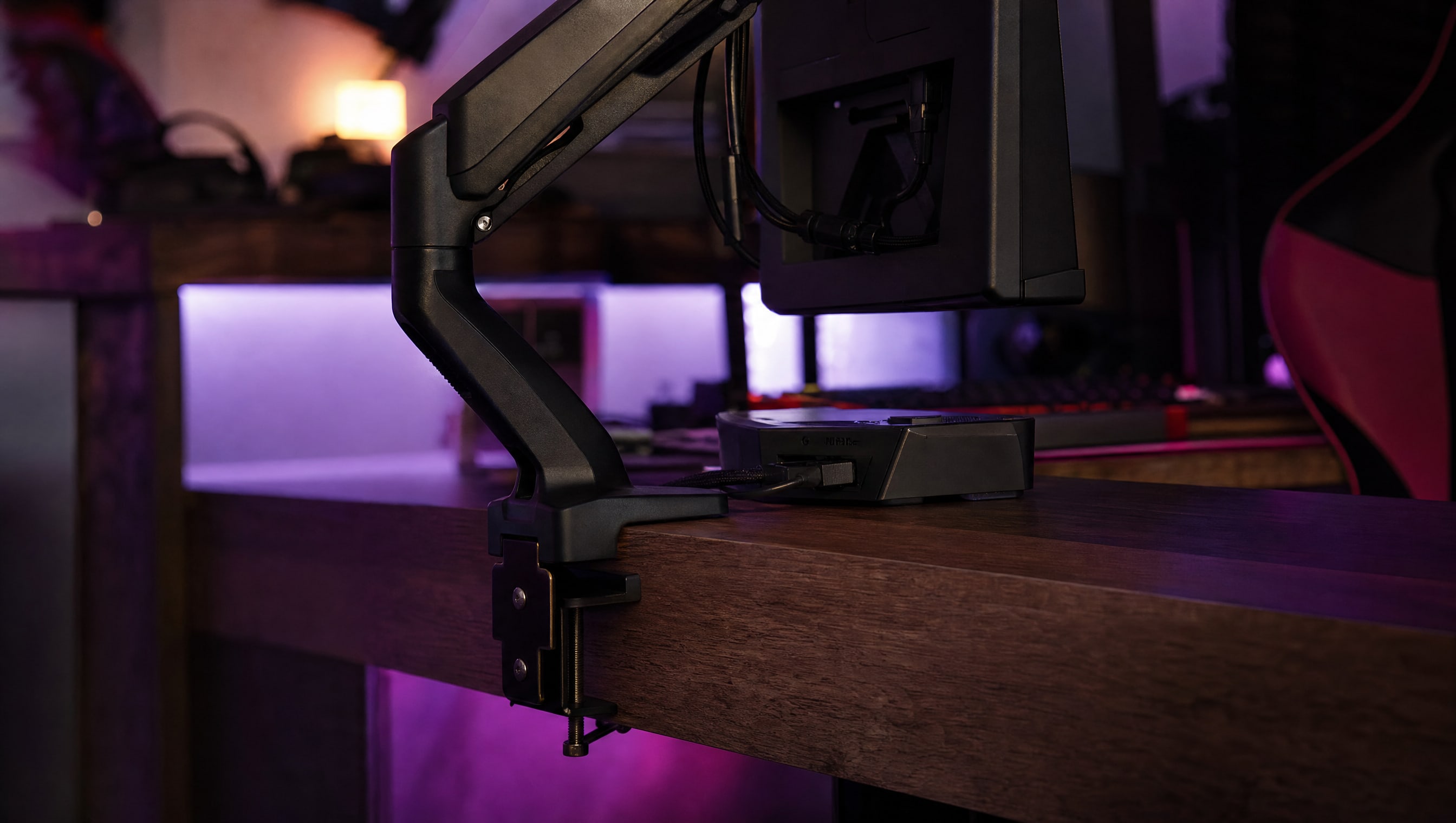 A monitor arm clamp biting the rear edge of a desk, the jaw and pad visible.